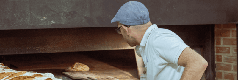 Baker taking off baked breads with shovel from the professional oven at the manufacturing